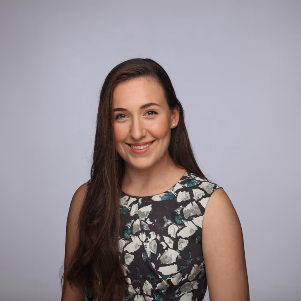 Smiling woman with long brown hair in a sleeveless floral dress against a plain gray background.