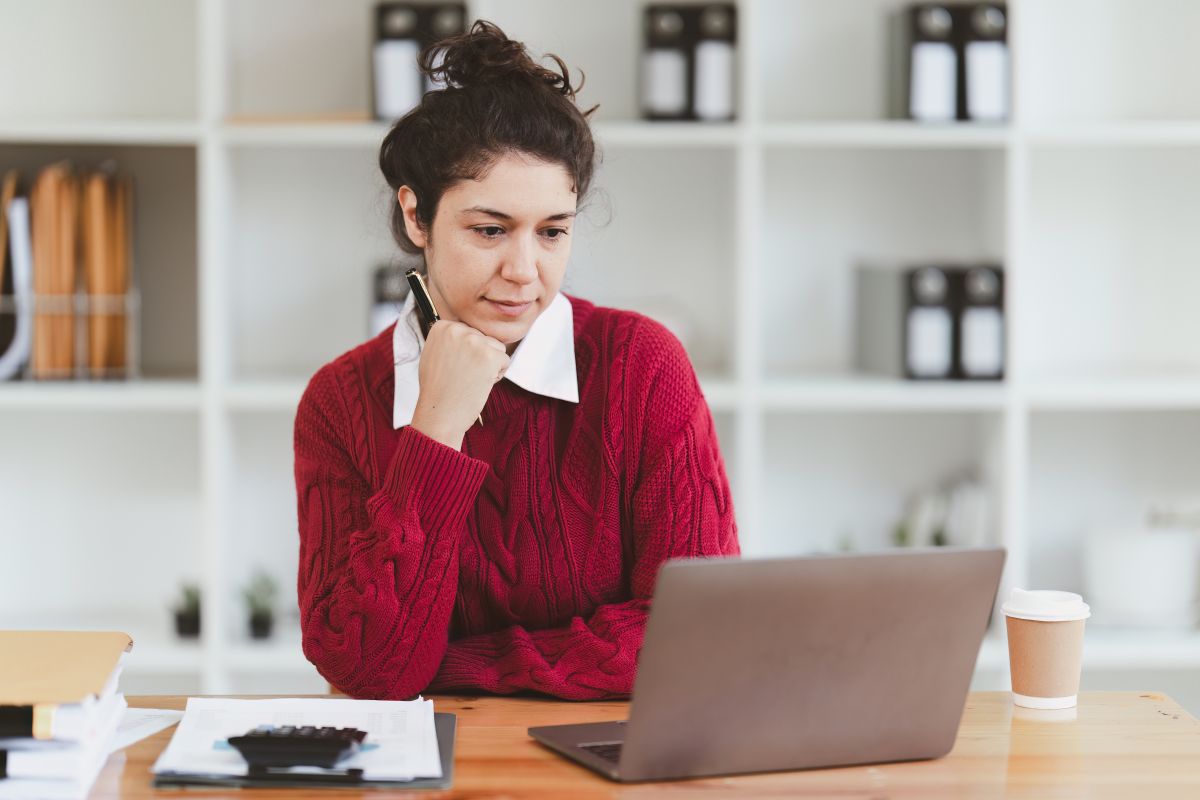 Woman in red sweater working on laptop with documents, calculator, and coffee cup on desk.