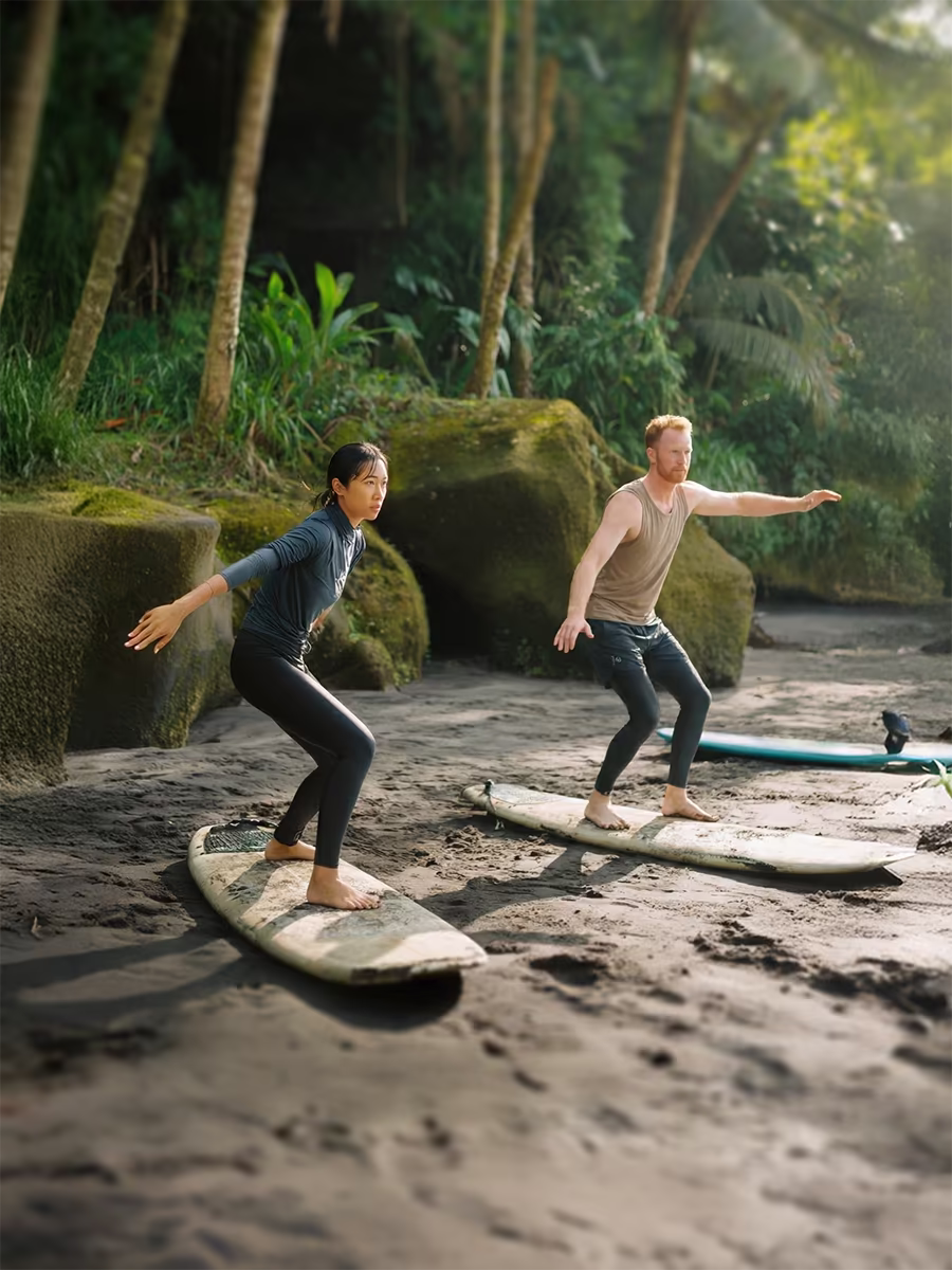 Two people practicing surfing stances on surfboards on a sandy beach with lush greenery and rocks in the background.