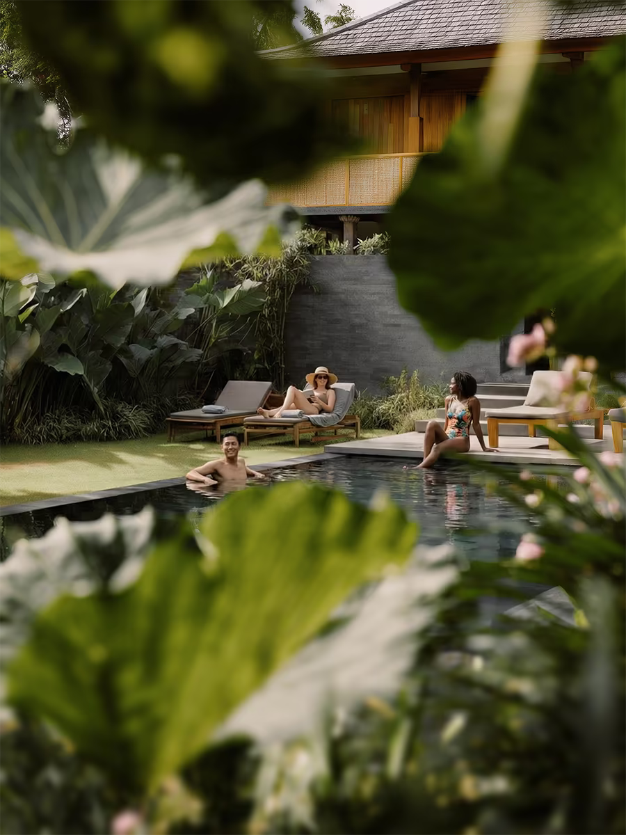 Three people relaxing by a pool surrounded by lush greenery and tropical plants, with a wooden villa in the background.