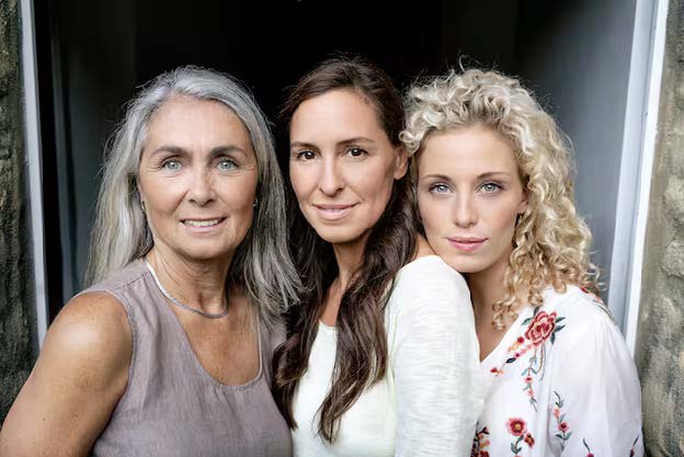 Drei Generationen Frauen lächeln nah zusammen, jede mit unterschiedlicher Haarfarbe und -stil.
