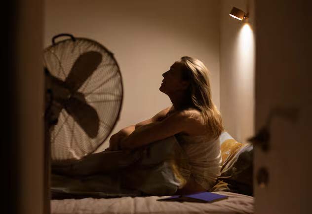 Eine Frau sitzt am Bett im dunklen Zimmer mit Ventilator und Buch auf dem Bett.