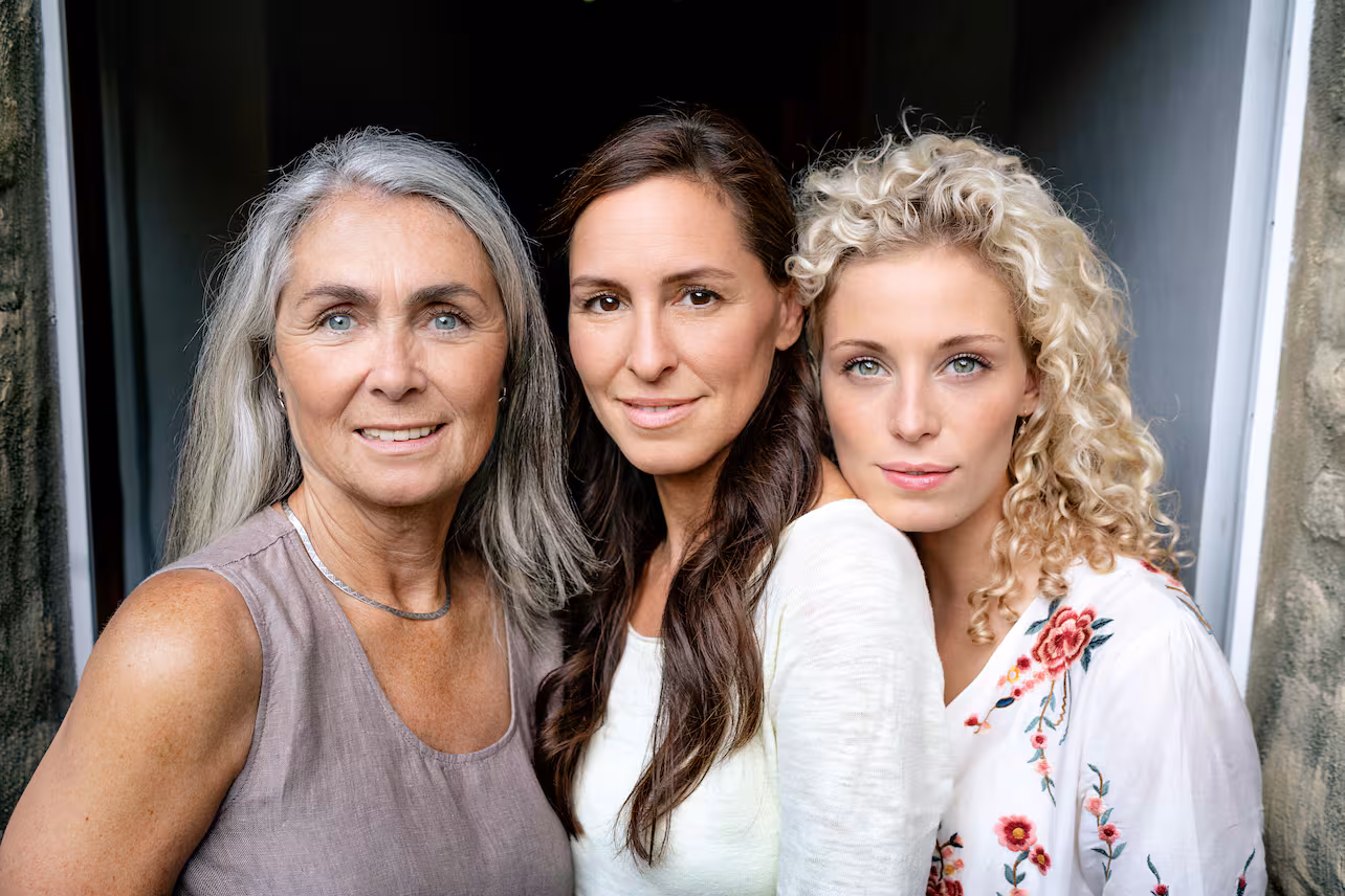 Drei Generationen Frauen lächeln nah zusammen, jede mit unterschiedlicher Haarfarbe und -stil.