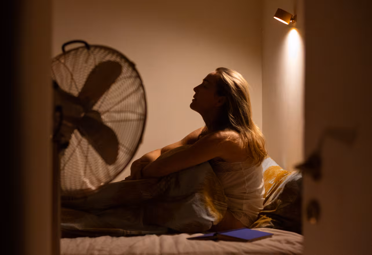 Eine Frau sitzt am Bett im dunklen Zimmer mit Ventilator und Buch auf dem Bett.