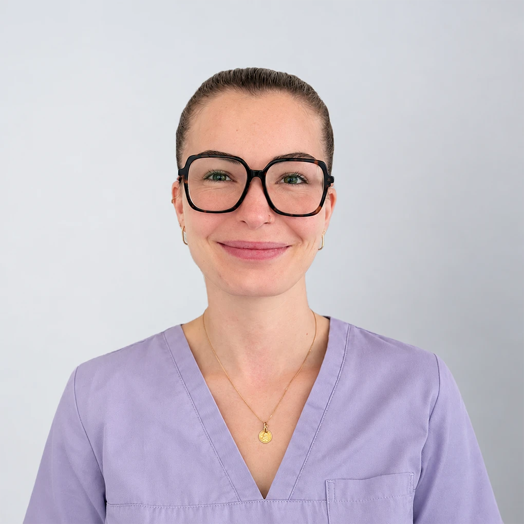 Smiling woman with short brown hair and glasses wearing a lavender medical scrub top against a plain gray background.