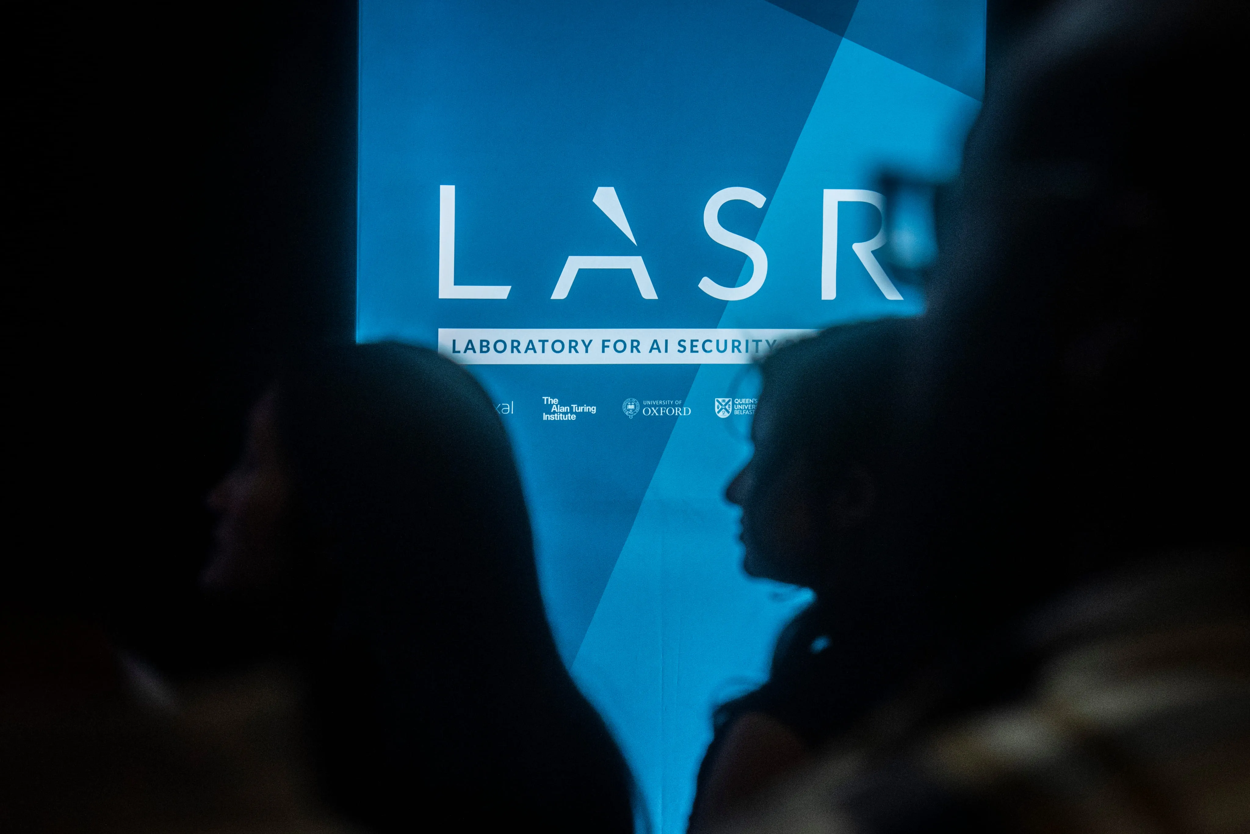 Silhouettes of people in front of a blue display reading 'LASR Laboratory for AI Security' with logos from partner institutions.