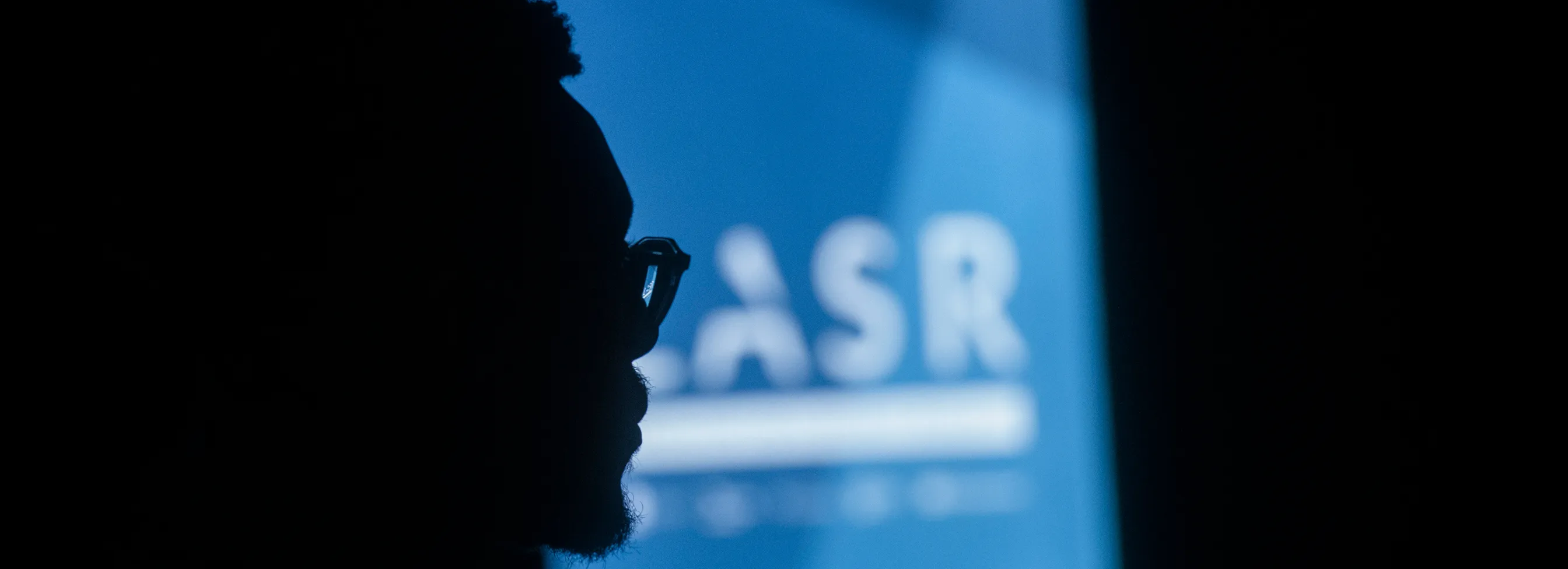 Silhouette of a person with glasses in front of a blue screen displaying the blurred text 'LASR'.