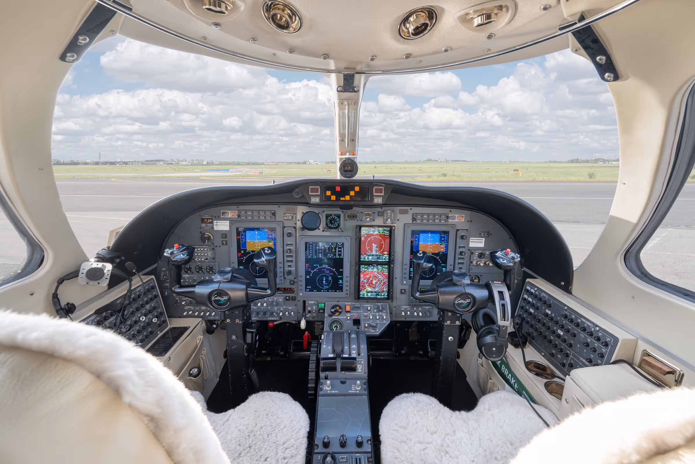 Interior view of a small aircraft cockpit with dual control yokes, digital instrument panels, and sheepskin-covered pilot seats.