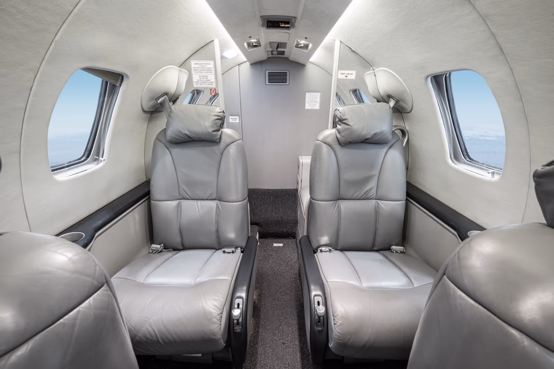 Interior of a small private jet with four gray leather seats, two on each side facing each other, and oval windows showing blue sky.