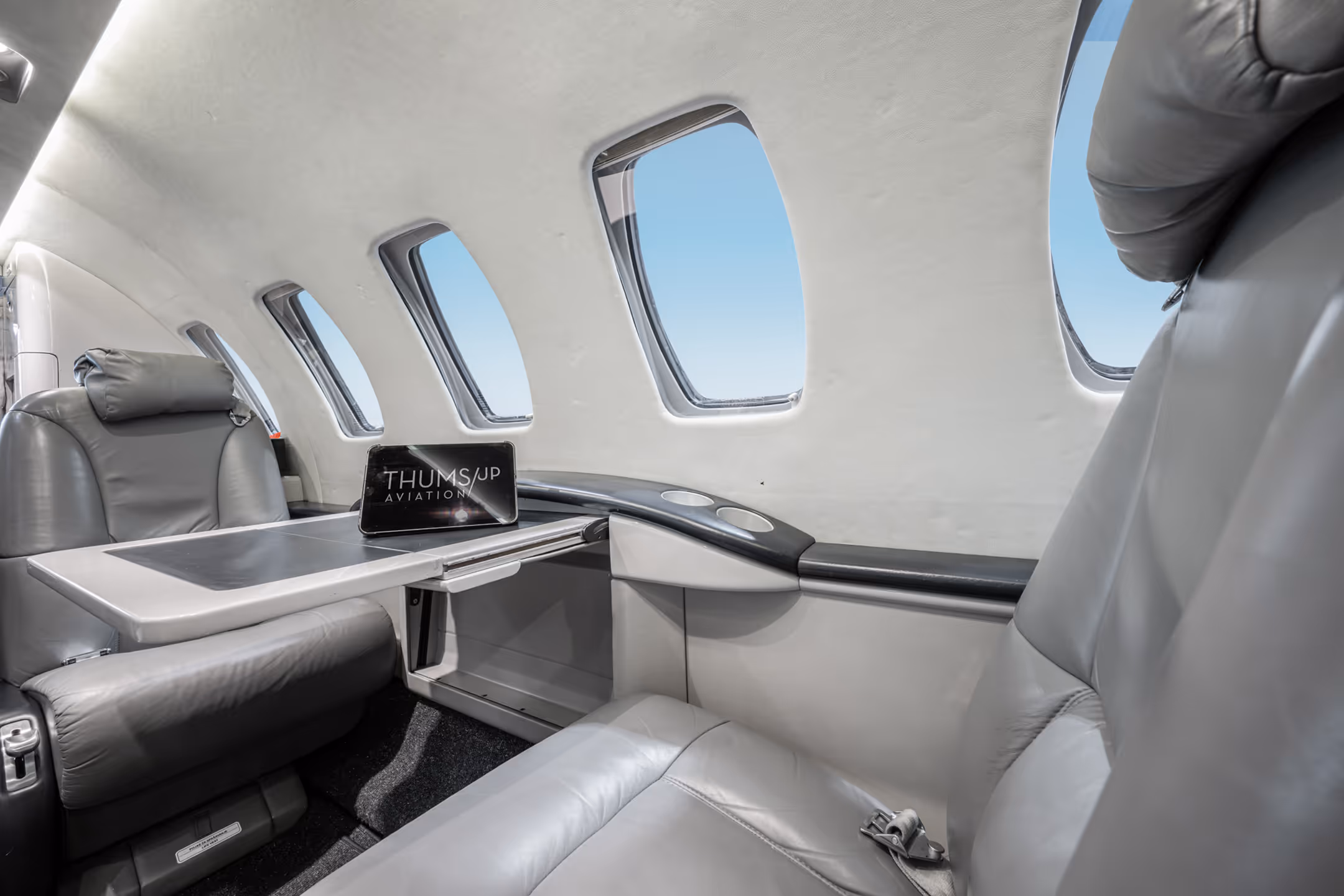 Interior of a private jet with gray leather seats, fold-out table, and five windows showing a blue sky.
