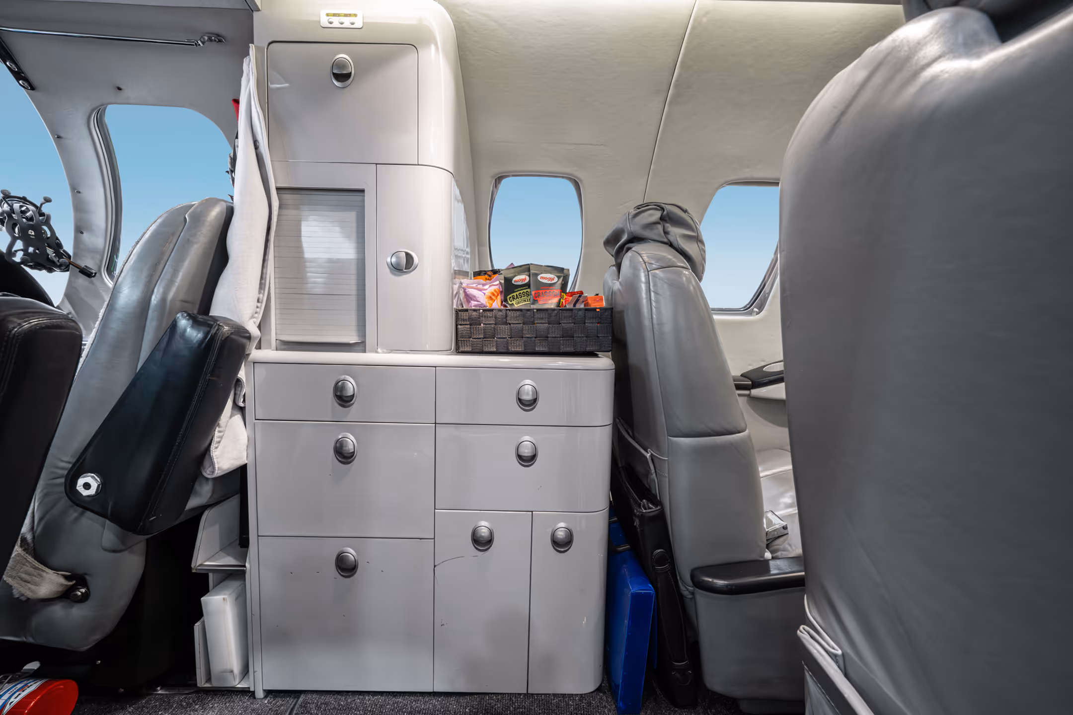 Interior of a private jet with gray leather seats and a storage cabinet holding a basket of snacks between the seats.