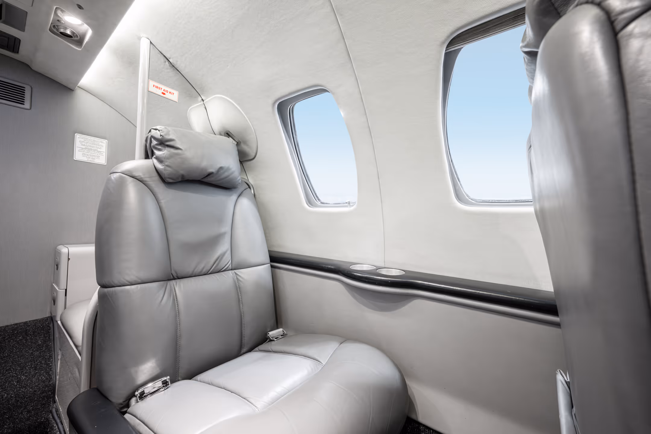 Gray leather passenger seat inside a private jet cabin next to two windows with blue sky outside.