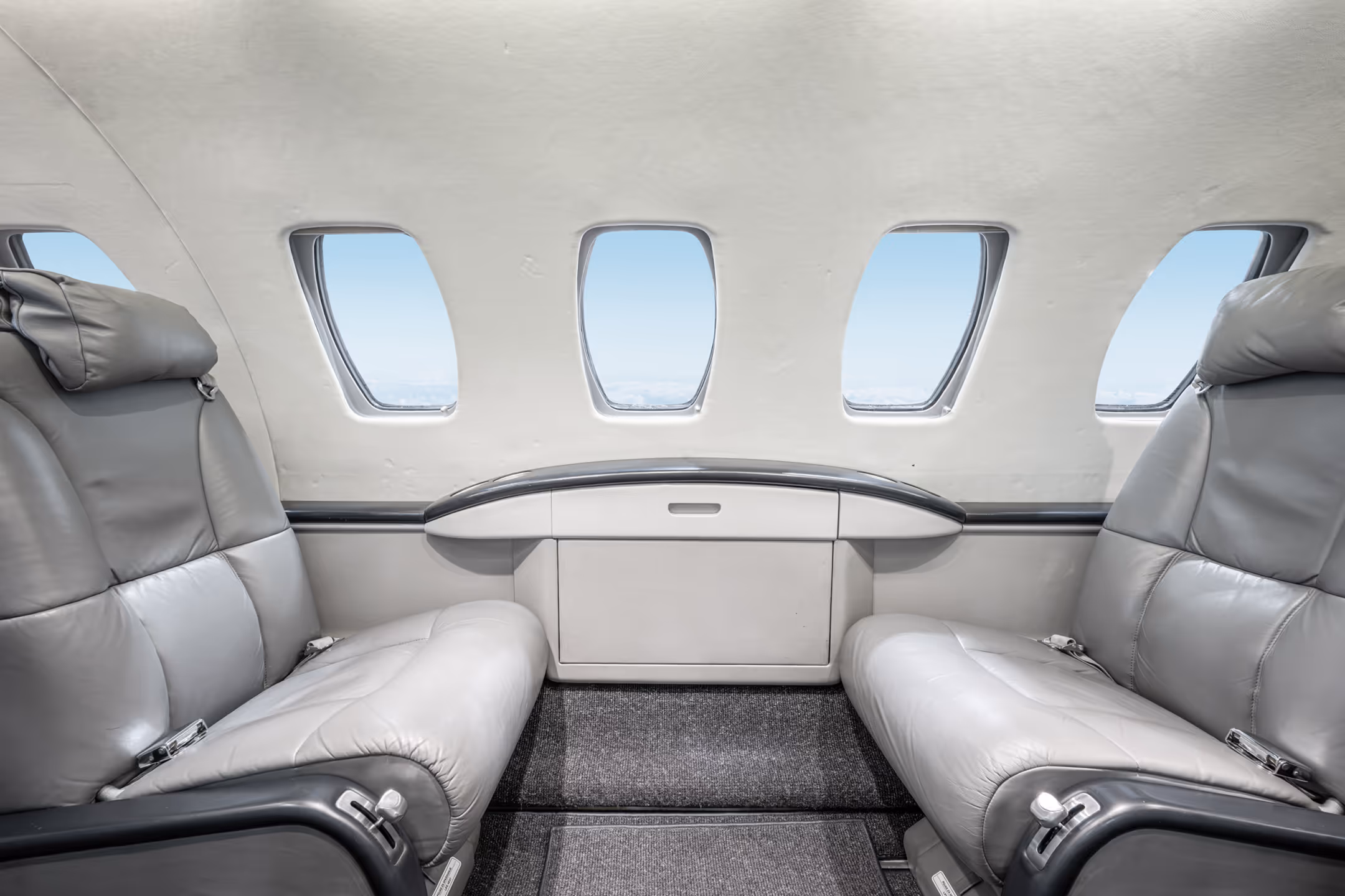 Interior of a small private jet with two facing gray leather seats and four windows showing a blue sky.