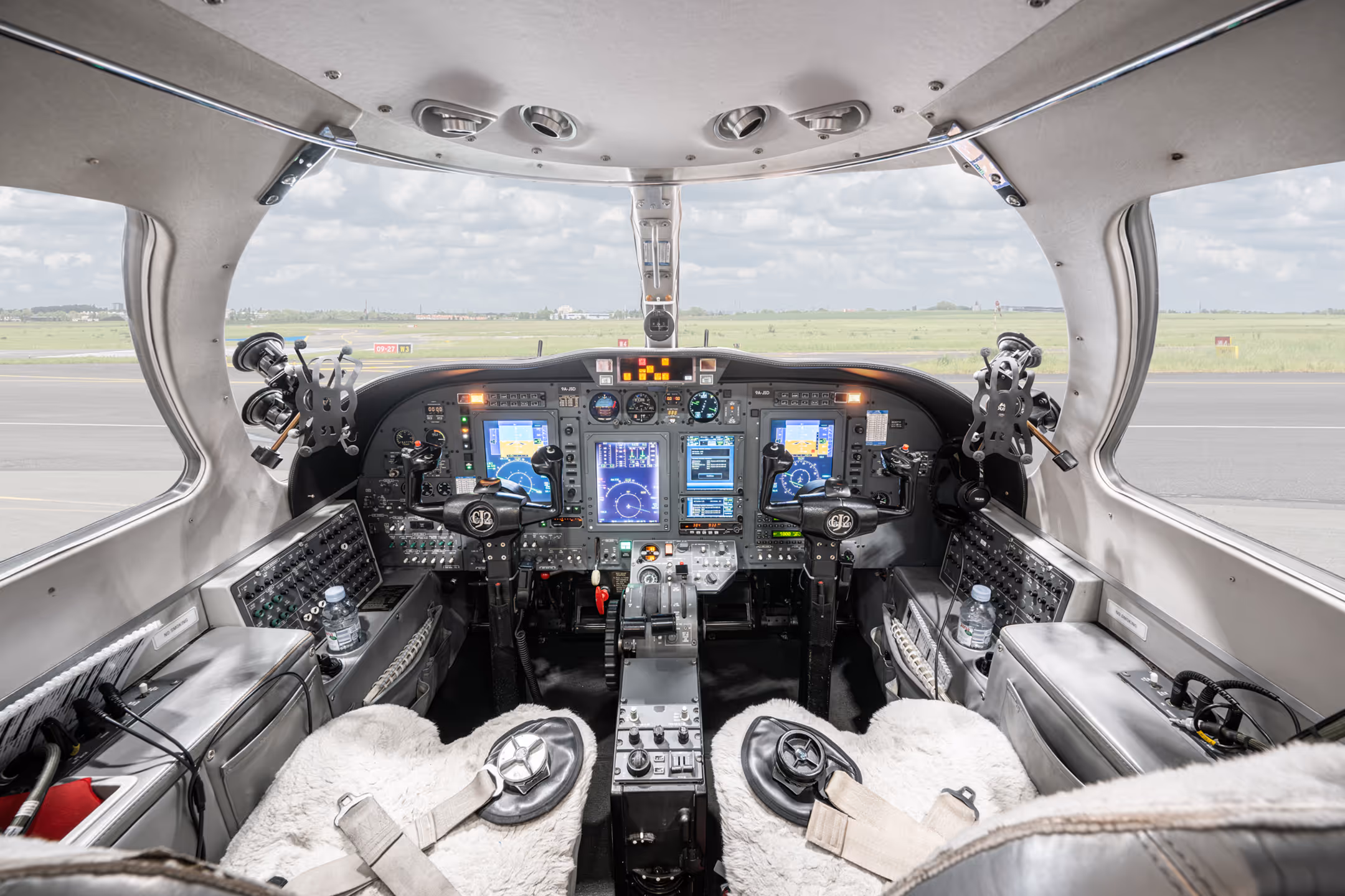 Cockpit interior of a small aircraft showing dual pilot seats with sheepskin covers, control yokes, multiple digital flight instrument displays, and a view of the runway and grassy field through the windshield.