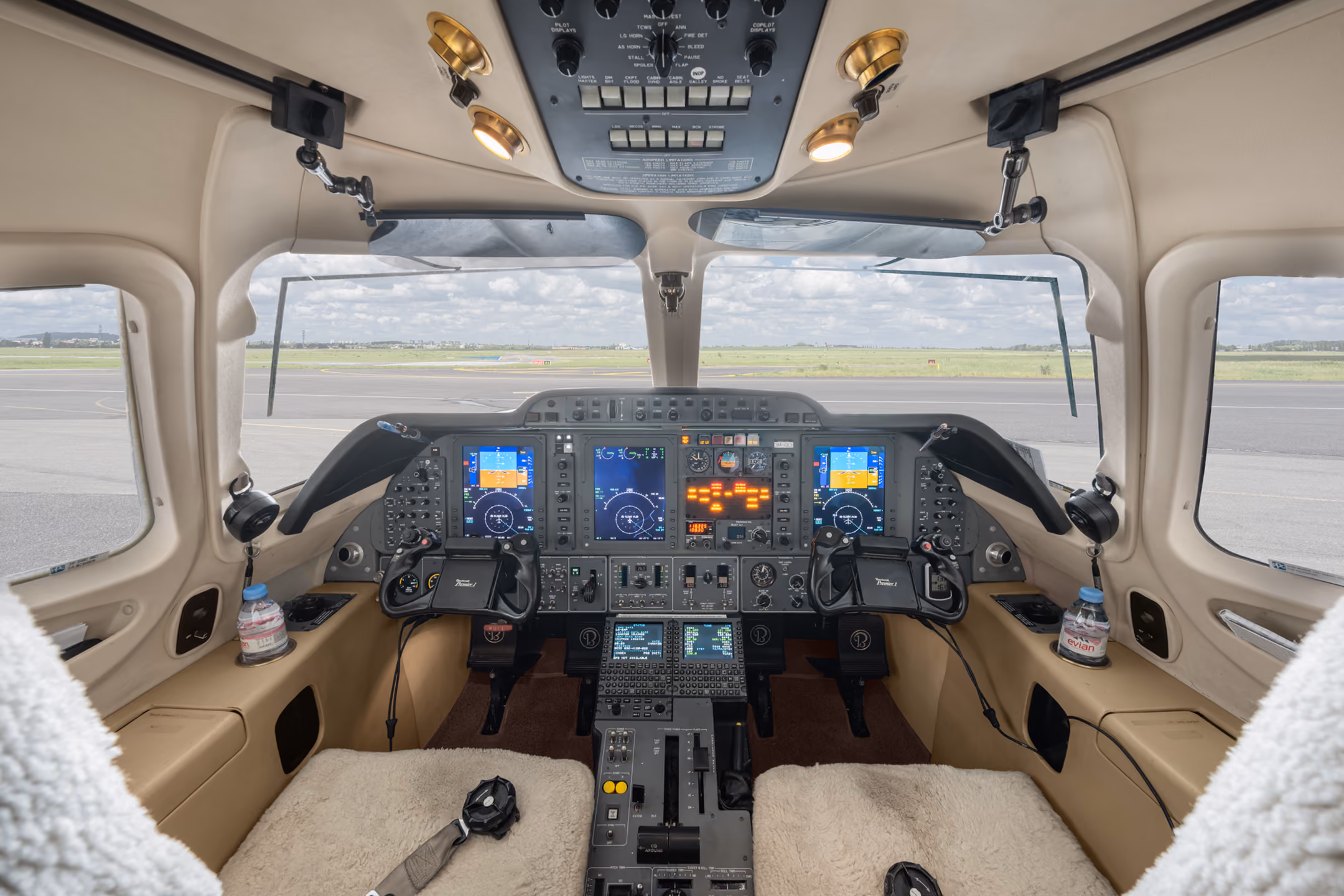 Cockpit interior of a small aircraft showing dual pilot seats with sheepskin covers, control yokes, multiple digital flight instrument displays, and a view of the runway and grassy field through the windshield.