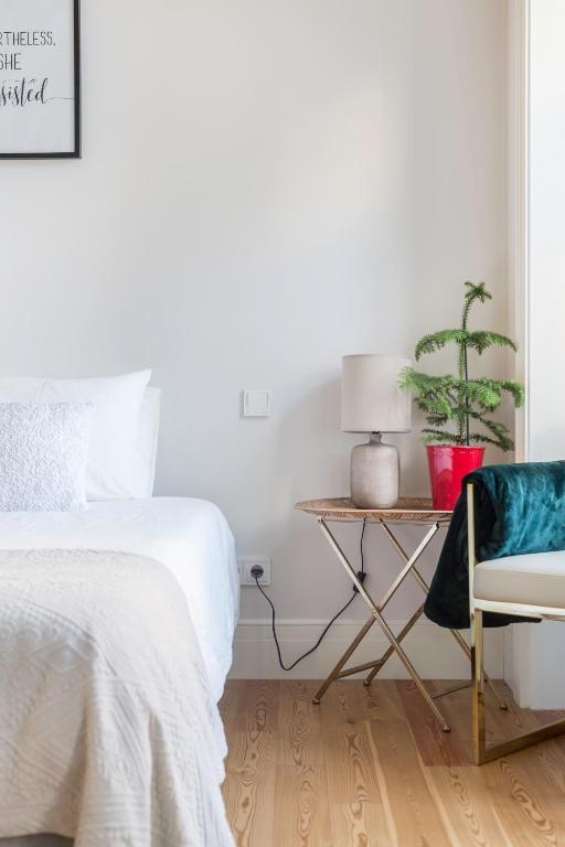 Minimalist bedroom corner with white bed, wooden floor, side table with lamp and potted plant.