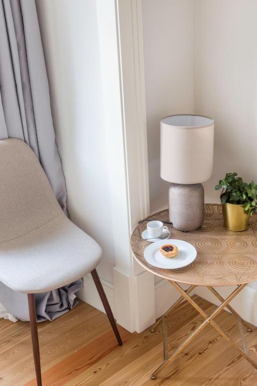 Beige chair next to a wooden round table with a lamp, a plant, cup, and plate with pastry.