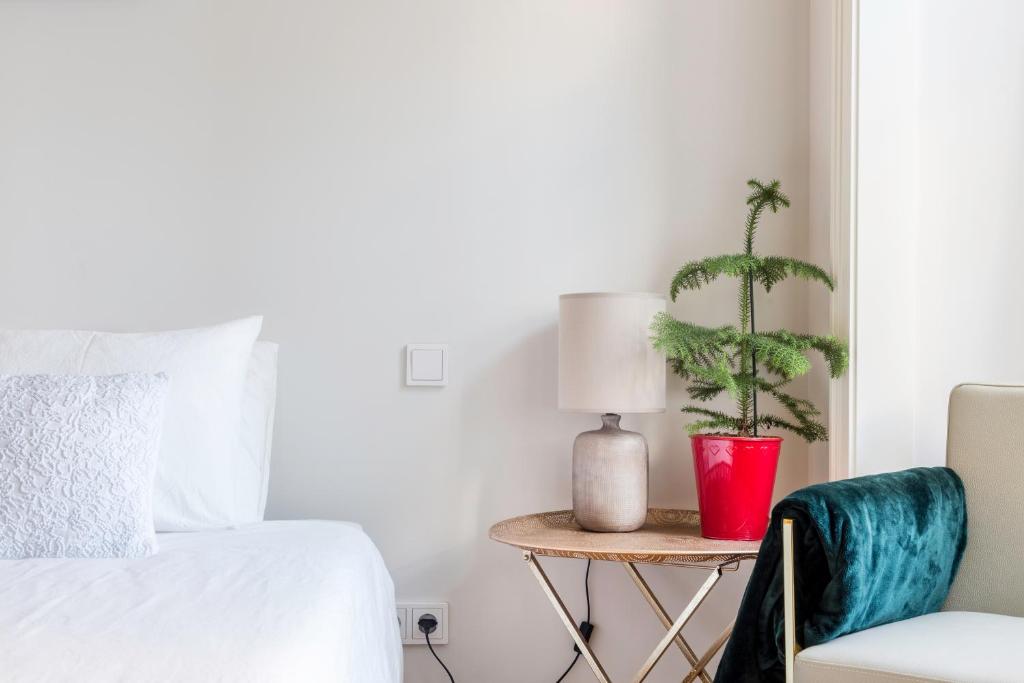 Minimalist bedroom corner with white bed, textured pillow, side table, lamp, small plant in red pot, and teal blanket.