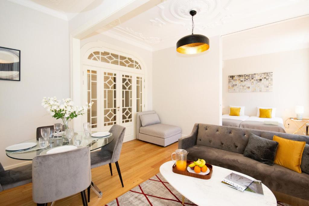 Bright living room with gray sofa, glass dining table, white doors, and a glimpse into a bedroom with yellow pillows.