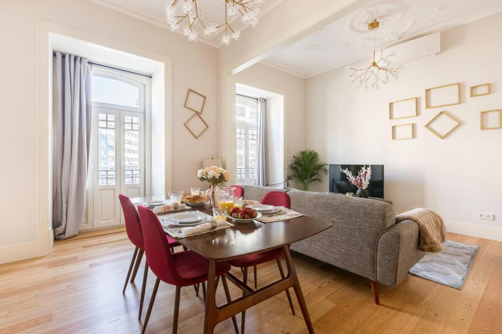Bright dining room with a wooden table, red chairs, gray sofa, wall decor, and large windows with curtains.