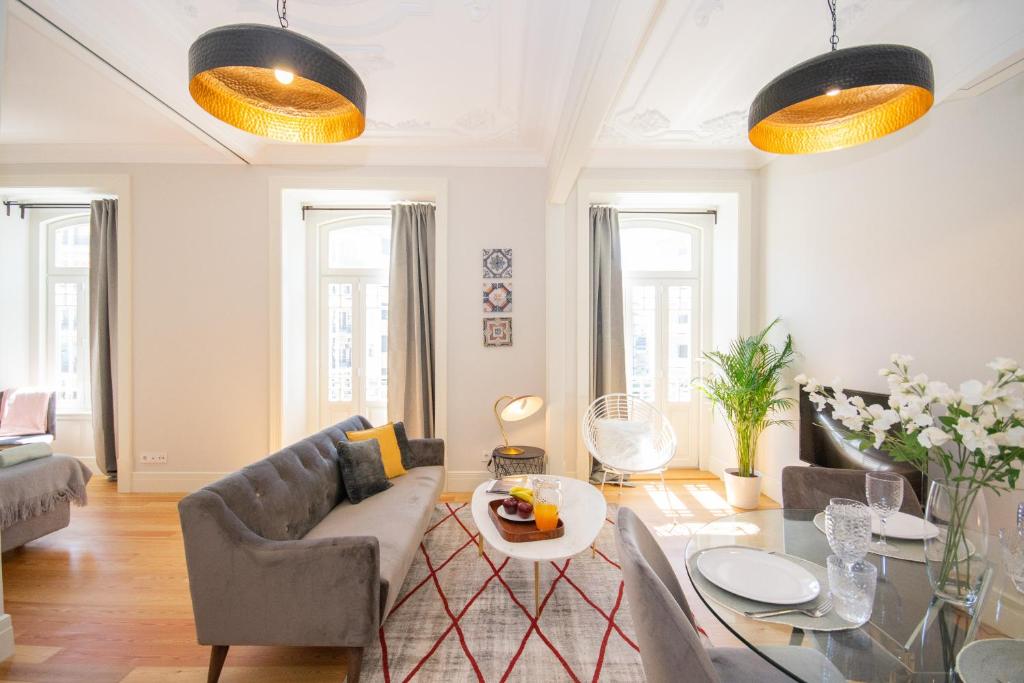 Bright living room with gray sofa, white chair, glass table set for two, plants, and large windows.