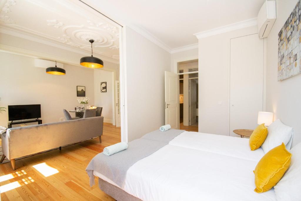 Bright bedroom with two beds, yellow pillows, and a view into a living room with gray sofa and dining table.