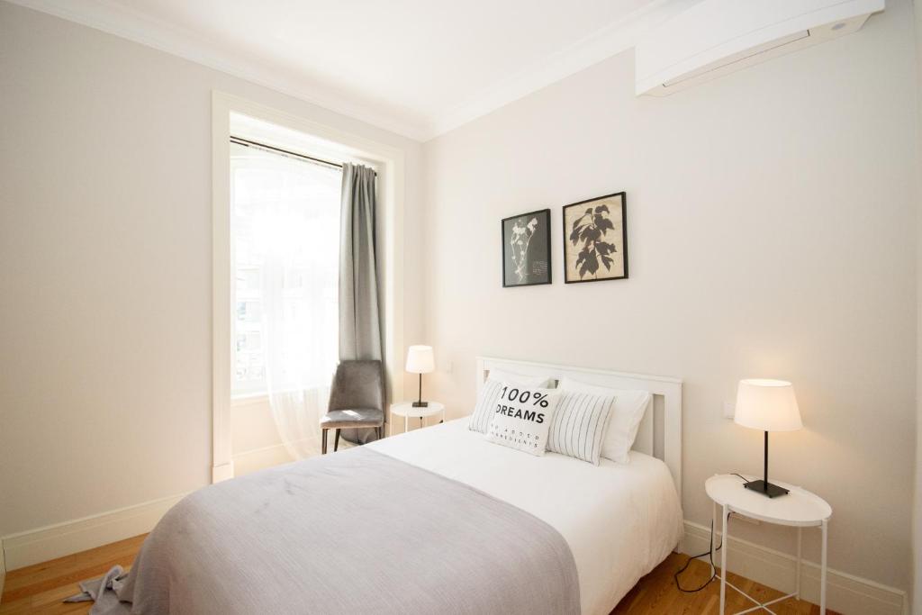 Bright bedroom with white bed, gray blanket, two side tables with lamps, and a chair near window.