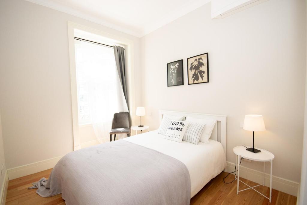 Minimalist bedroom with a white bed, gray blanket, two pillows, side tables with lamps, and framed leaf art.