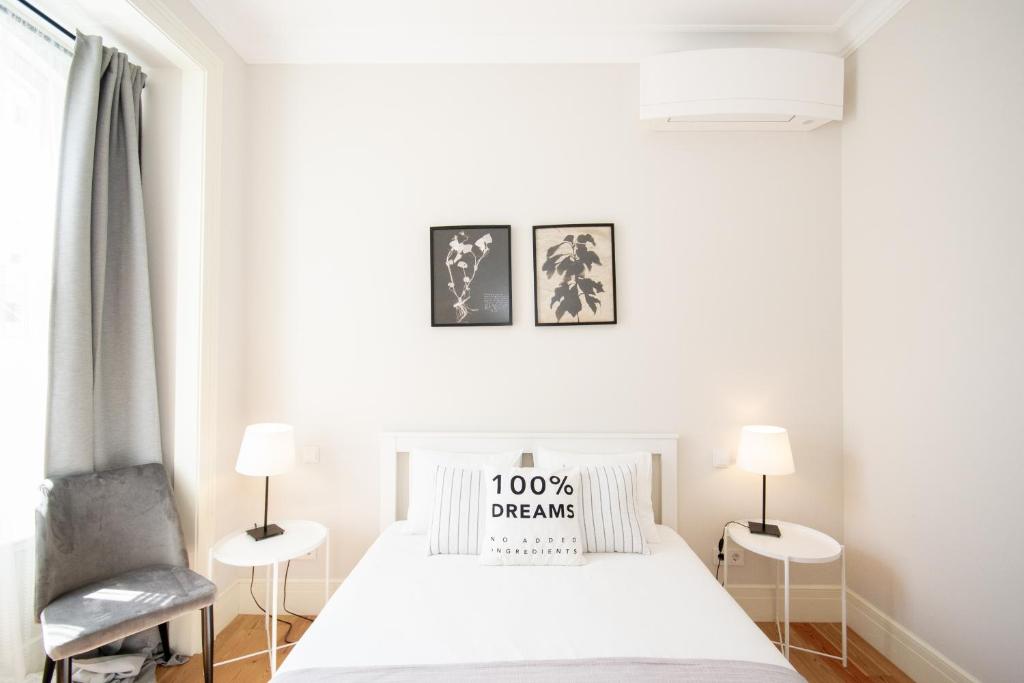 Minimalist bedroom with white bed, pillows including one reading '100% DREAMS', side tables, lamps, and framed plant art.