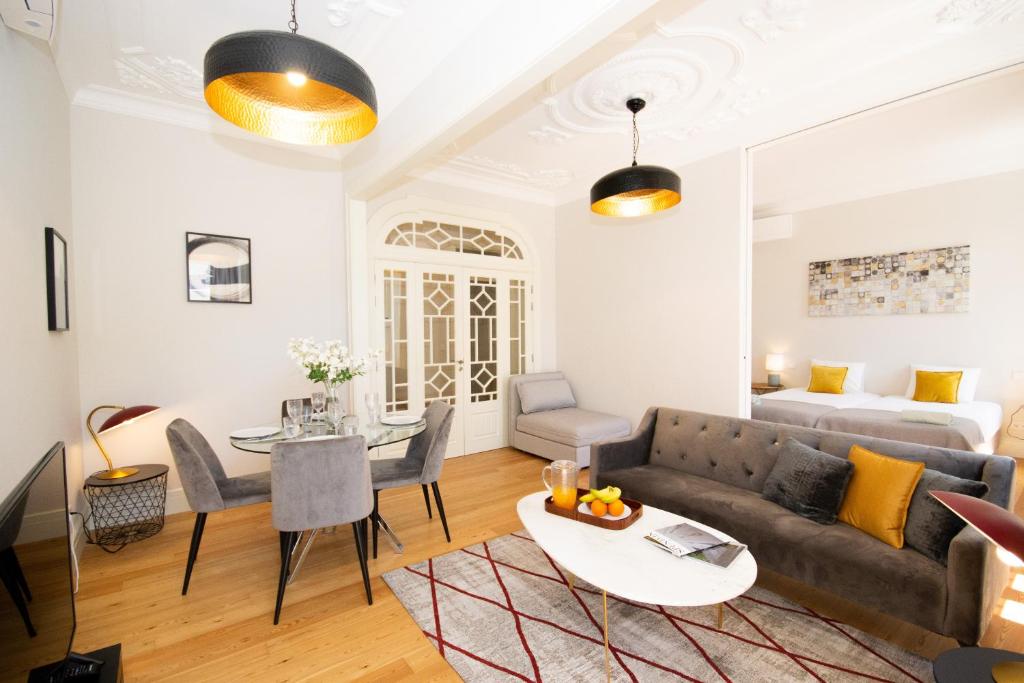 Modern living room with gray sofa, glass dining table with chairs, and a bedroom with two beds in background.