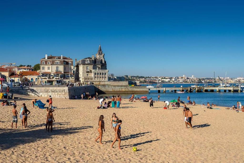 People enjoying a sunny day on a sandy beach near historic buildings by calm blue water.