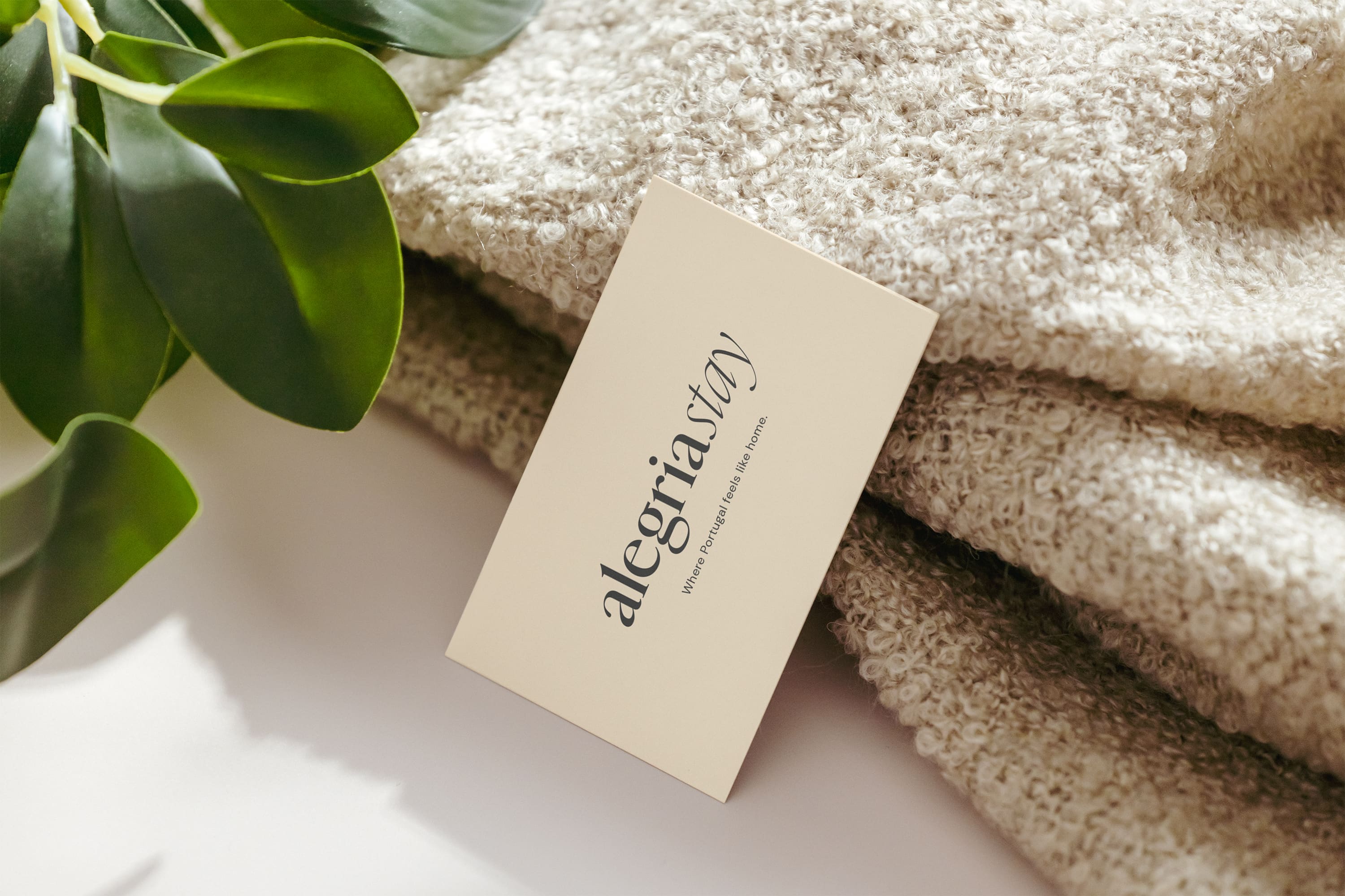 Business card with 'alegriastay' text on textured beige fabric next to green leaves.