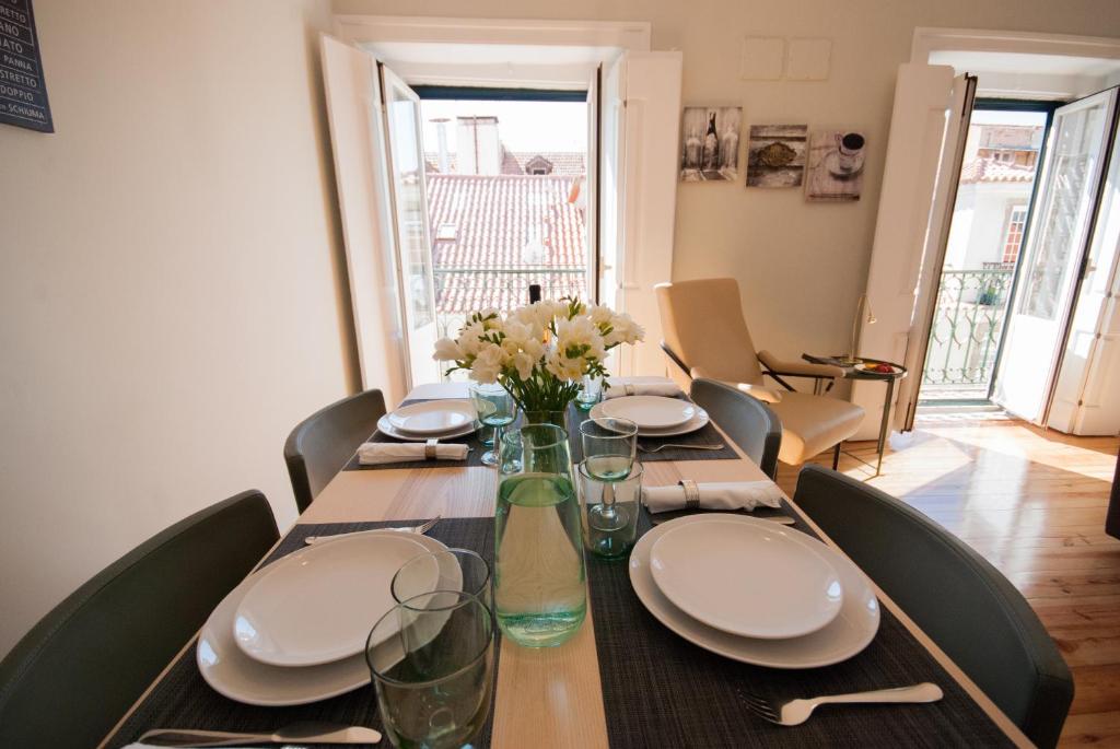 Dining table set with plates, glasses, and a vase of white flowers near open windows with city view.
