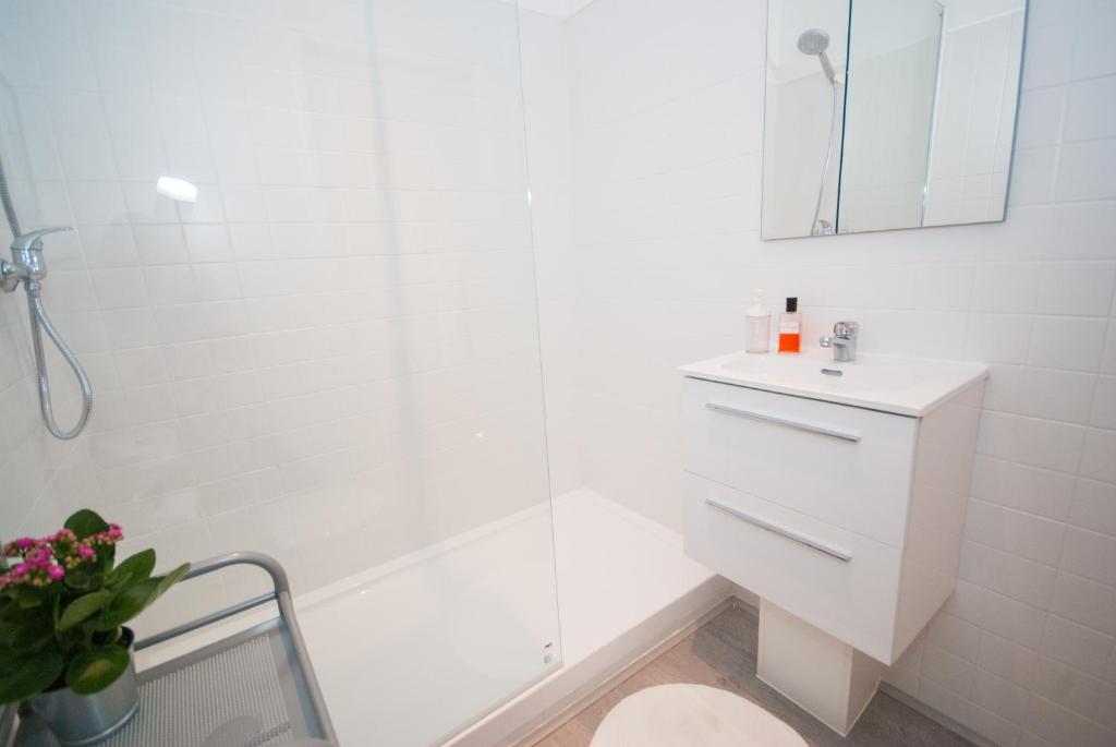 Bright white bathroom with glass shower, small vanity with mirror, and potted plant on metal shelf.