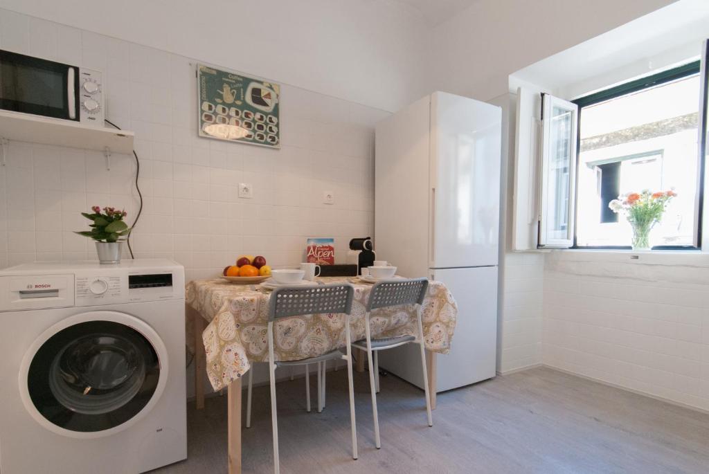 Compact kitchen with washing machine, microwave, fridge, table with fruit, cereal, and two chairs by window.