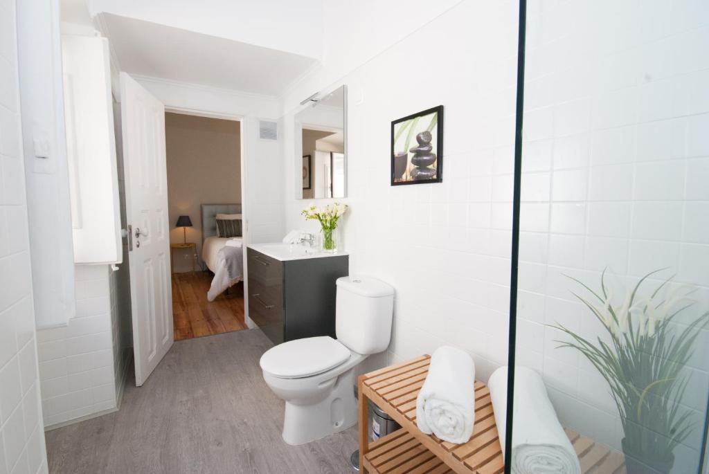 Bright bathroom with white tiles, toilet, wooden shelf with towels, and doorway to bedroom.