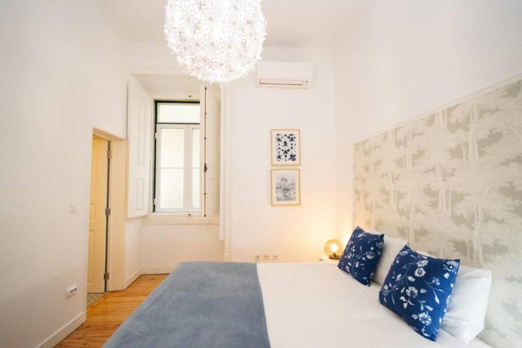 Bright bedroom with blue floral pillows, white bedding, patterned wall, window, and a hanging light fixture.
