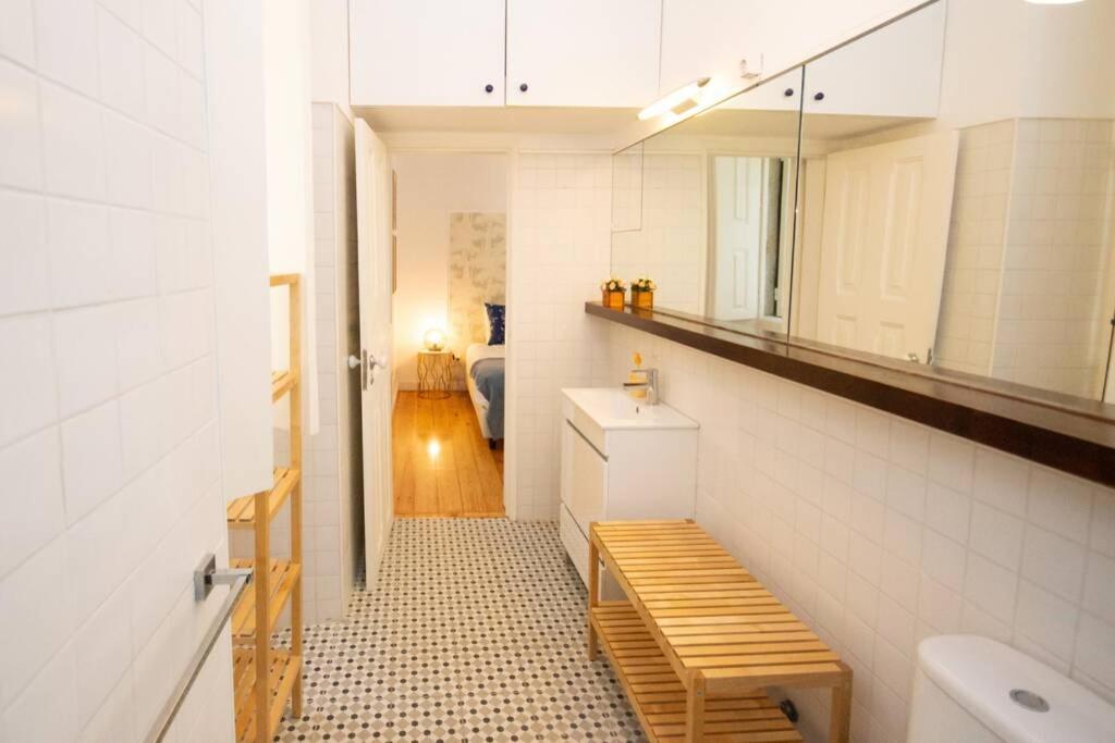 Narrow white-tiled bathroom with wooden bench, vanity, mirror, and view into a bedroom with wooden floor.