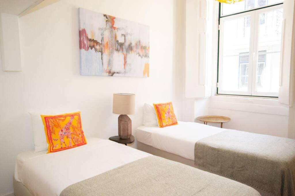 Bright room with two single beds, white bedding, gray throws, and orange patterned cushions by a sunlight window.