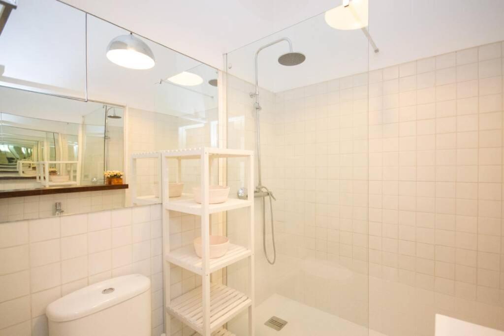 Bright white bathroom with a glass shower, overhead showerhead, shelving unit, and toilet.