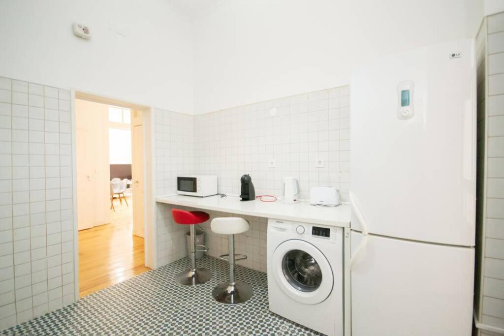 Bright kitchen corner with white fridge, washing machine, microwave, kettle, toaster, and two stools.