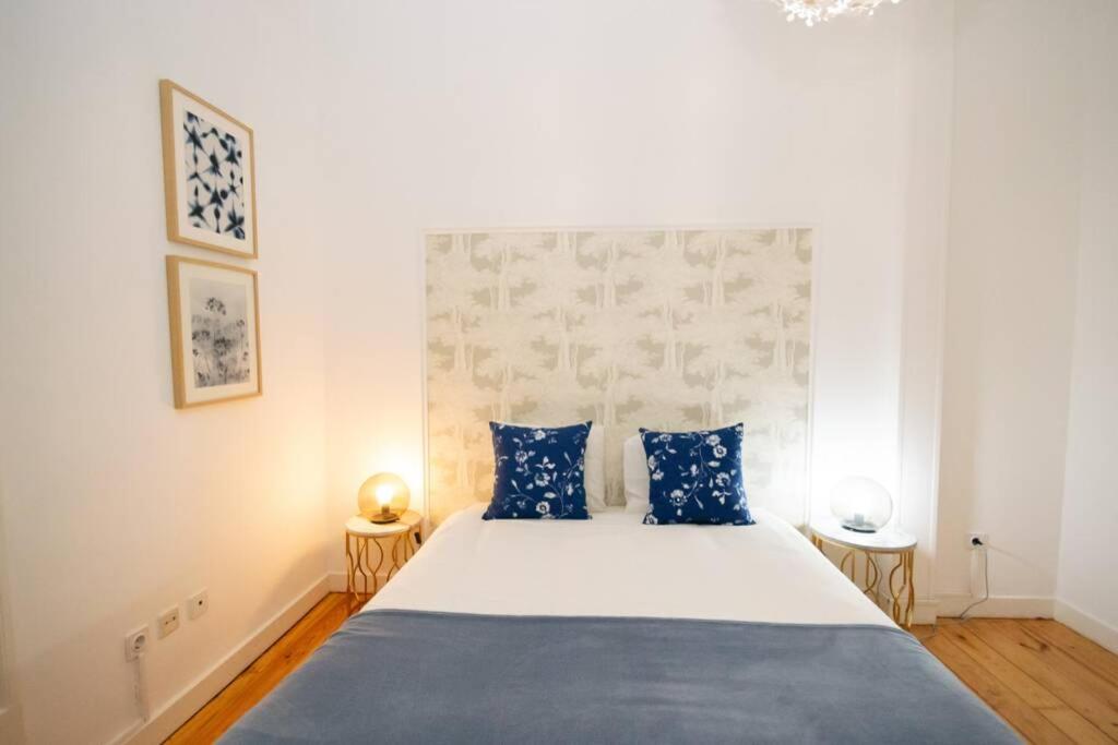Neatly made bed with white and blue bedding, floral pillows, two bedside tables with round lamps in a minimalist room.