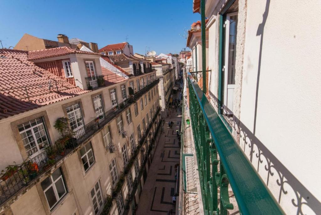 View down a narrow street with beige buildings and tile roofs from a balcony with green railing.
