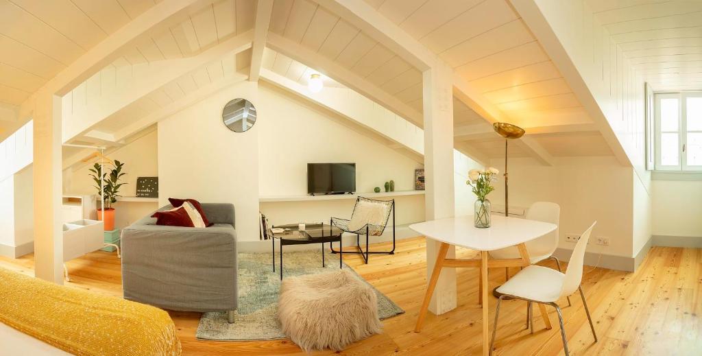 Bright attic living space with gray sofa, glass table, white chairs, wooden floor, and slanted ceiling beams.