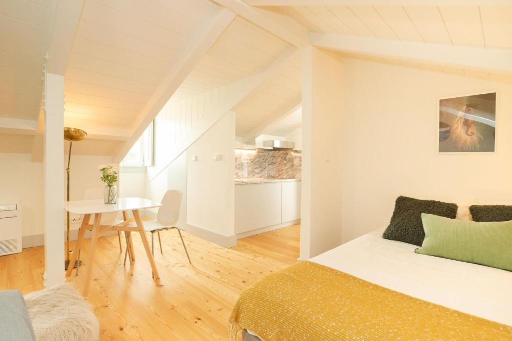 Bright attic room with wooden floor, white table and chairs, small kitchen, and bed with green pillows.