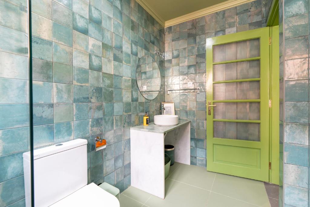 Bathroom with green tiled walls, white toilet, round mirror, green door, and white sink on marble countertop.