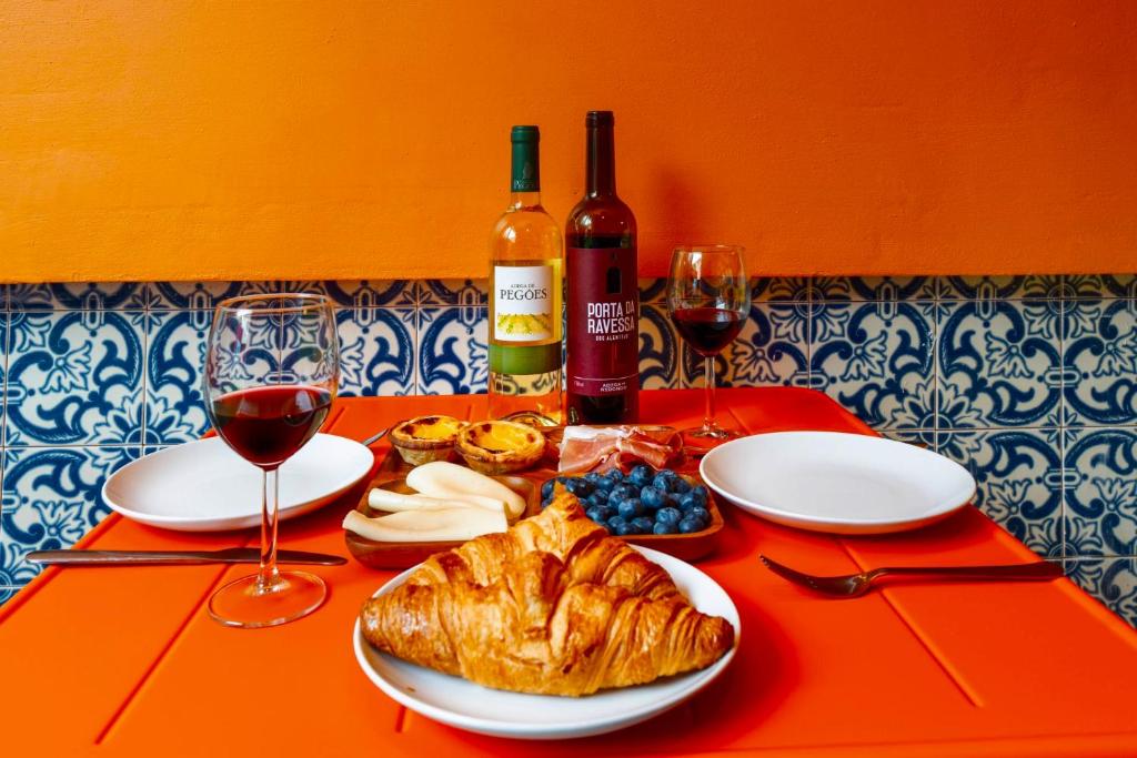 Table with croissant, blueberries, cheeses, meats, two wine glasses with red wine, and two bottles against orange wall.