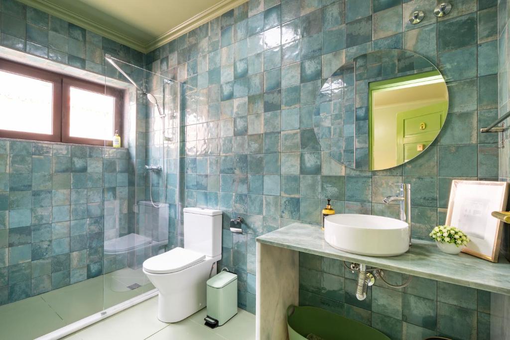 Bathroom with teal tiled walls, white toilet, round mirror, vessel sink, and glass shower enclosure.