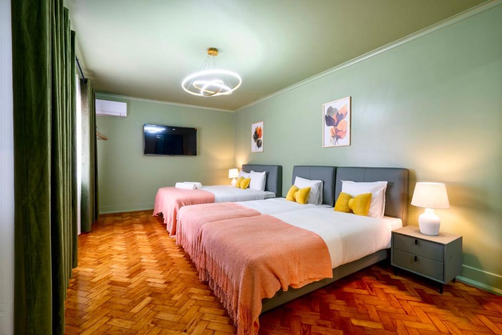 Hotel room with two beds, pink throws, yellow pillows, green walls, wooden floor, TV, and nightstand lamps.