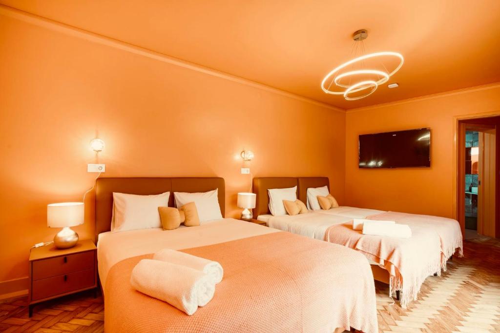 Hotel room with two double beds, orange walls, rolled towels, bedside lamps, and a wall-mounted TV.